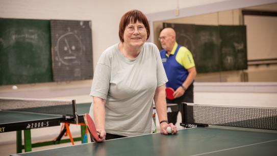 Tafeltennisster Josée aan een tafeltennistafel met op de achtergrond man die ook aan het spelen is, klik voor een vergroting